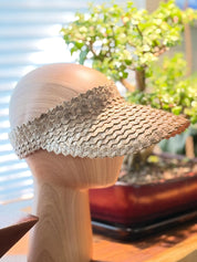 A wooden head wearing a woven sun visor is placed on a wooden surface, with a potted plant in the background.