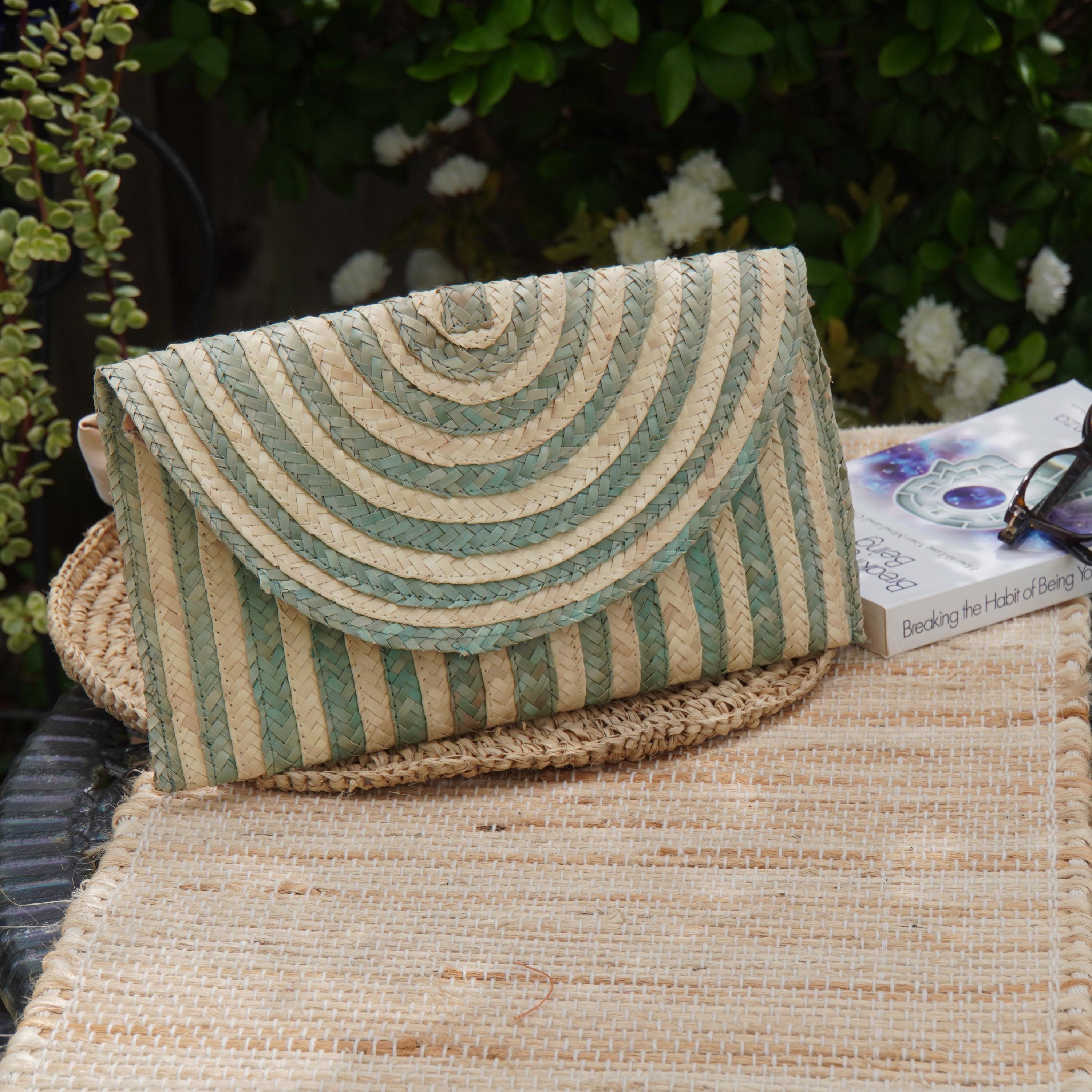 A woven purse with a green and white striped pattern rests on a woven placemat, accompanied by a book and a pair of sunglasses.