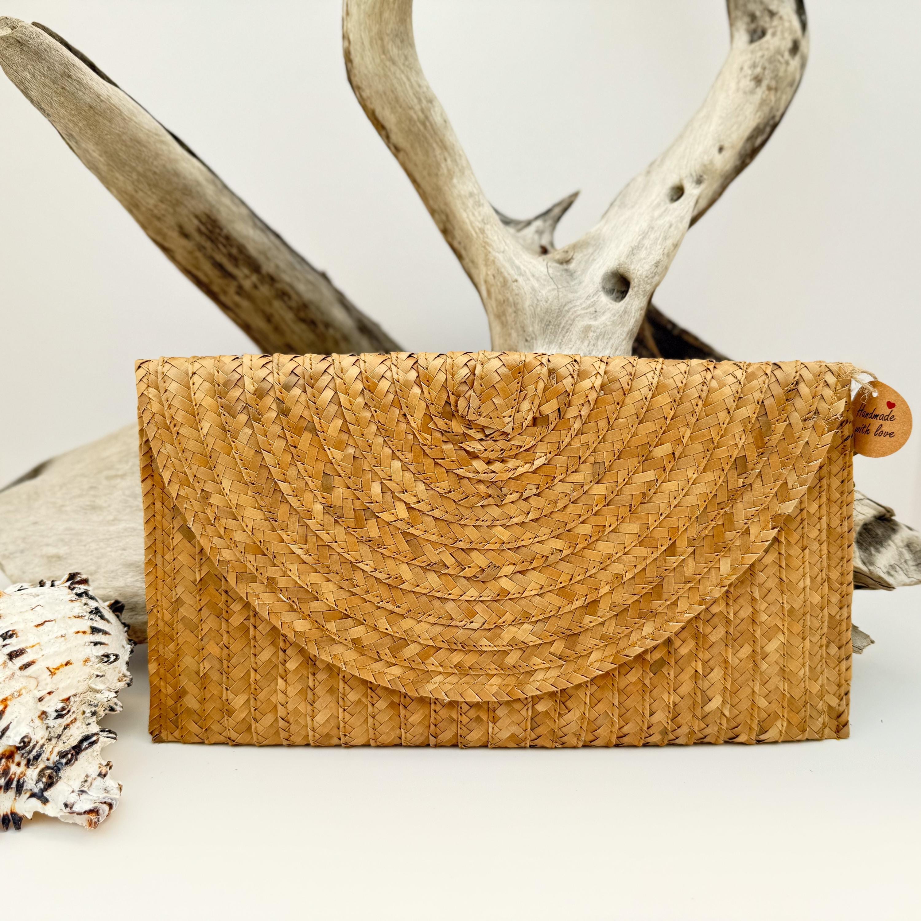A woven straw purse with a textured, woven pattern sits on a white surface, with a wooden branch and a small tag visible in the background.