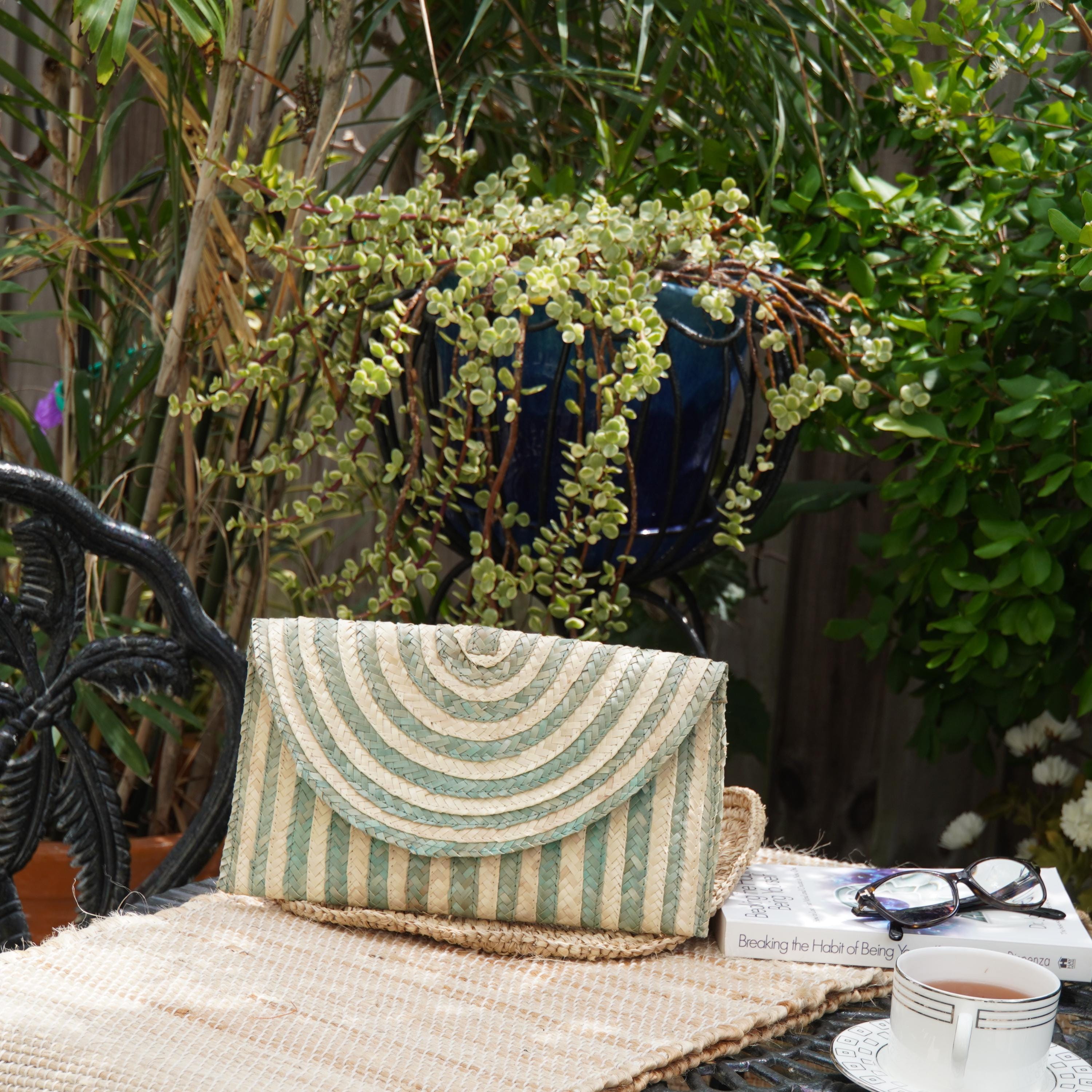 A striped purse rests on a woven placemat on a table, with a book and a cup of tea nearby. The table is set against a backdrop of lush greenery, including potted plants and a wooden fence.