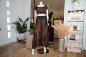 a mannequin wearing a black and orange patterned dress, standing on a round rug in a room with various decorative elements such as potted plants and a glass display case.