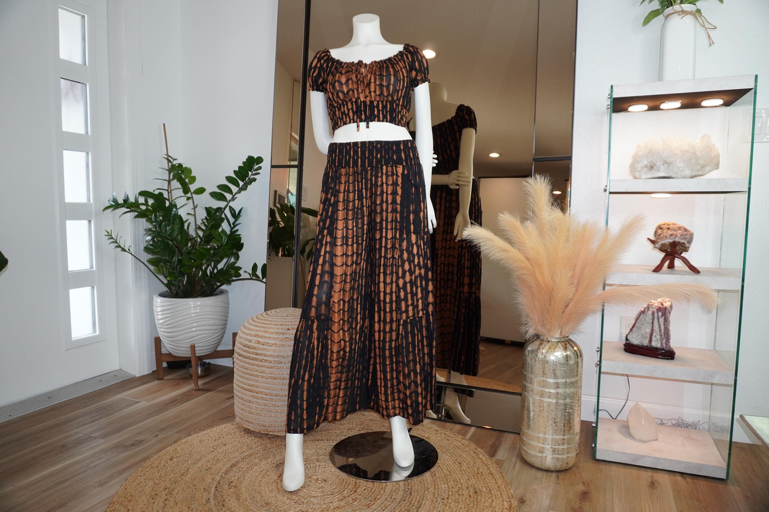 a mannequin wearing a black and orange patterned dress, standing on a round rug in a room with various decorative elements such as potted plants and a glass display case.