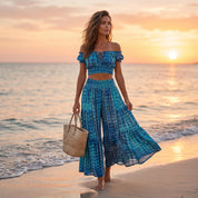 A woman in a blue and white patterned dress stands on a beach, holding a woven basket, with the sun setting in the background.