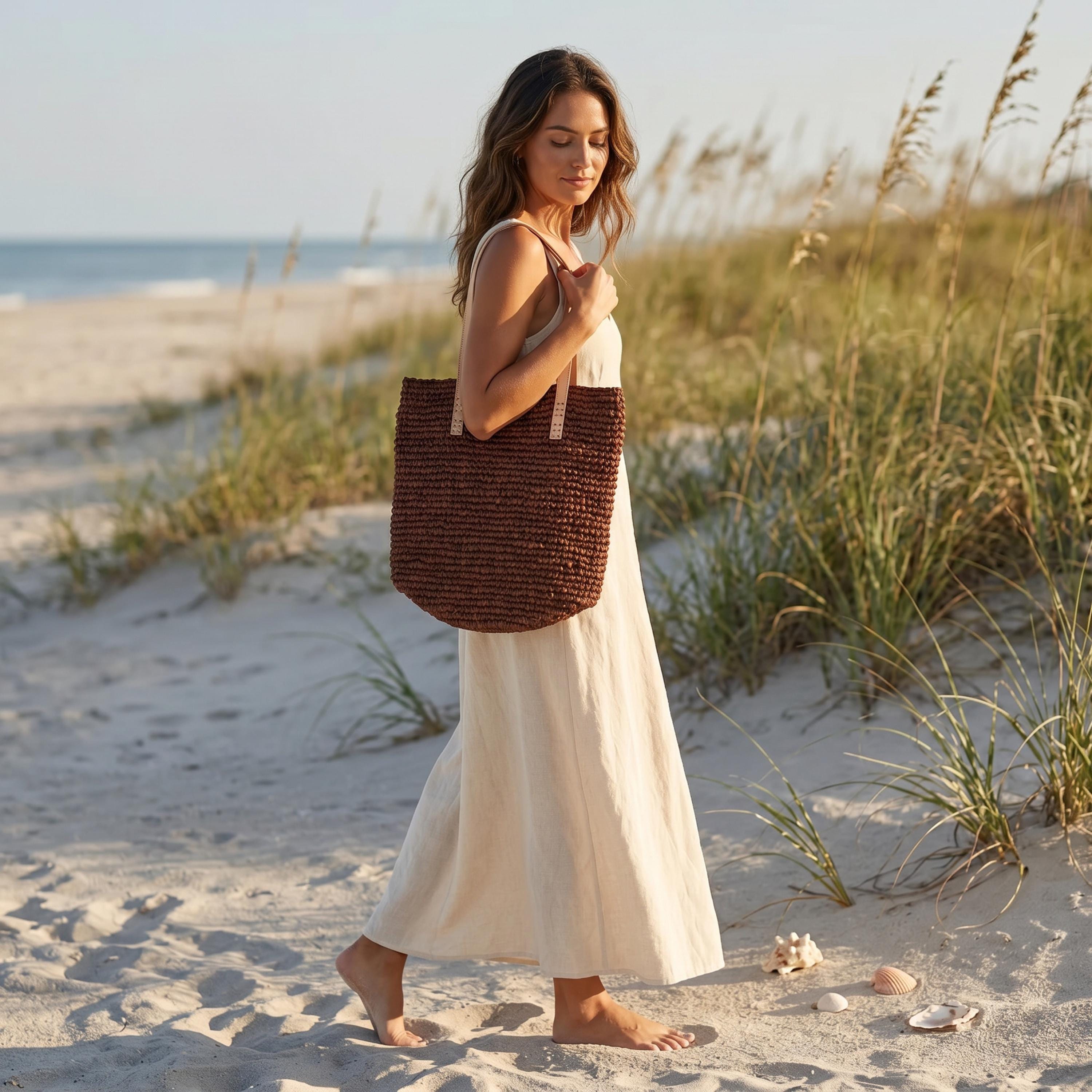 A woman in a white dress walks along a sandy beach, carrying a brown woven bag.