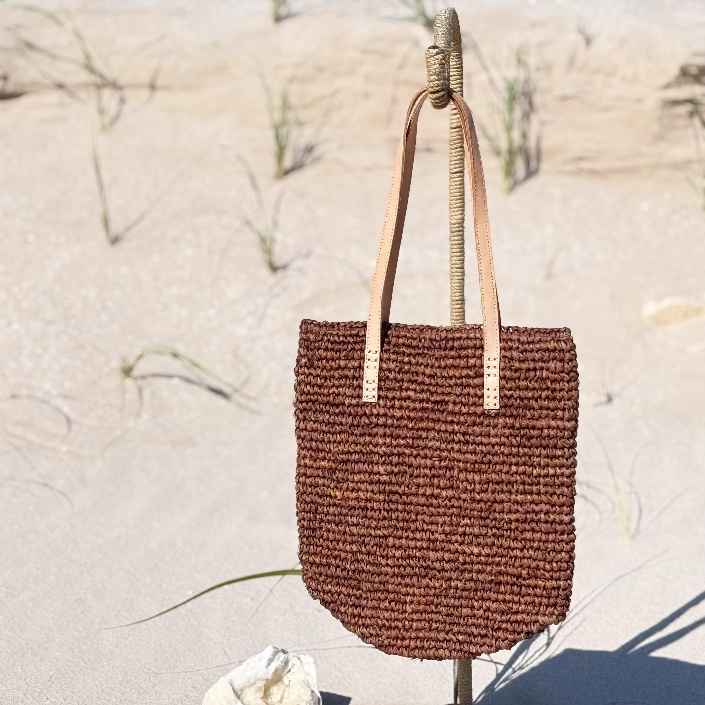 A brown woven handbag is hanging on a stick in a sandy beach setting.