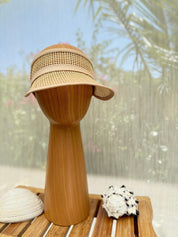 A straw hat with a woven pattern is displayed on a wooden stand, accompanied by a seashell and a small rock, against a blurred background of greenery.