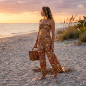 A woman in a floral print dress stands on a beach, holding a woven basket, with the ocean and sunset in the background.