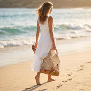 A woman in a white dress walks along a sandy beach, carrying a woven bag filled with various items.