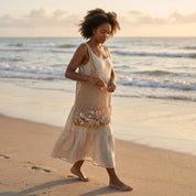 A woman in a beige dress walks along the beach, carrying a woven bag filled with seashells.