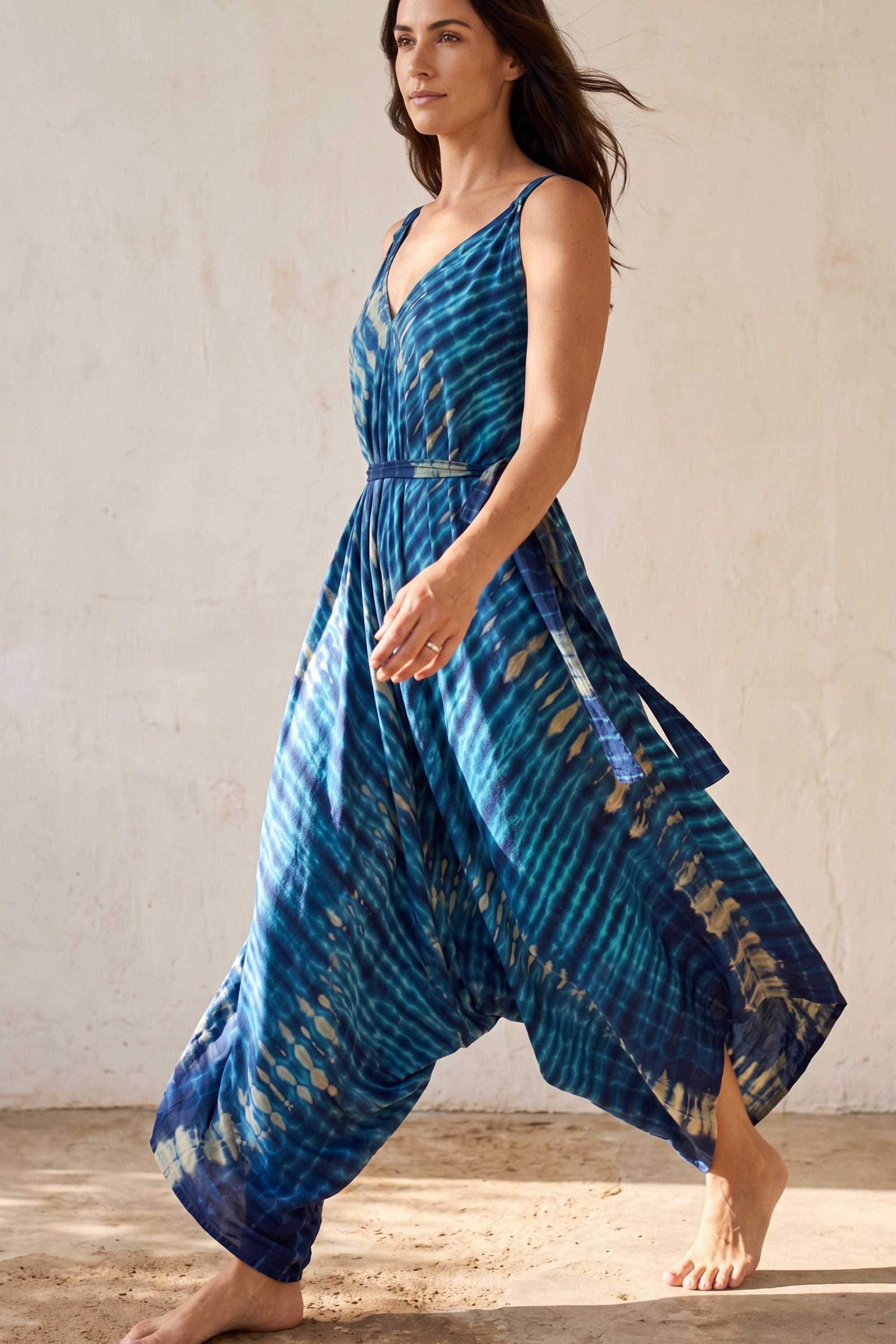 A woman wearing a blue and white tie-dye dress is walking barefoot in front of a plain white wall.