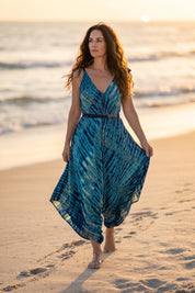 A woman in a blue and white tie-dye dress walks along a sandy beach during sunset, with the ocean visible in the background.