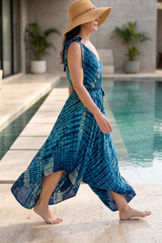 A woman wearing a blue and white tie-dye dress and a wide-brimmed hat walks along a tiled walkway near a pool.