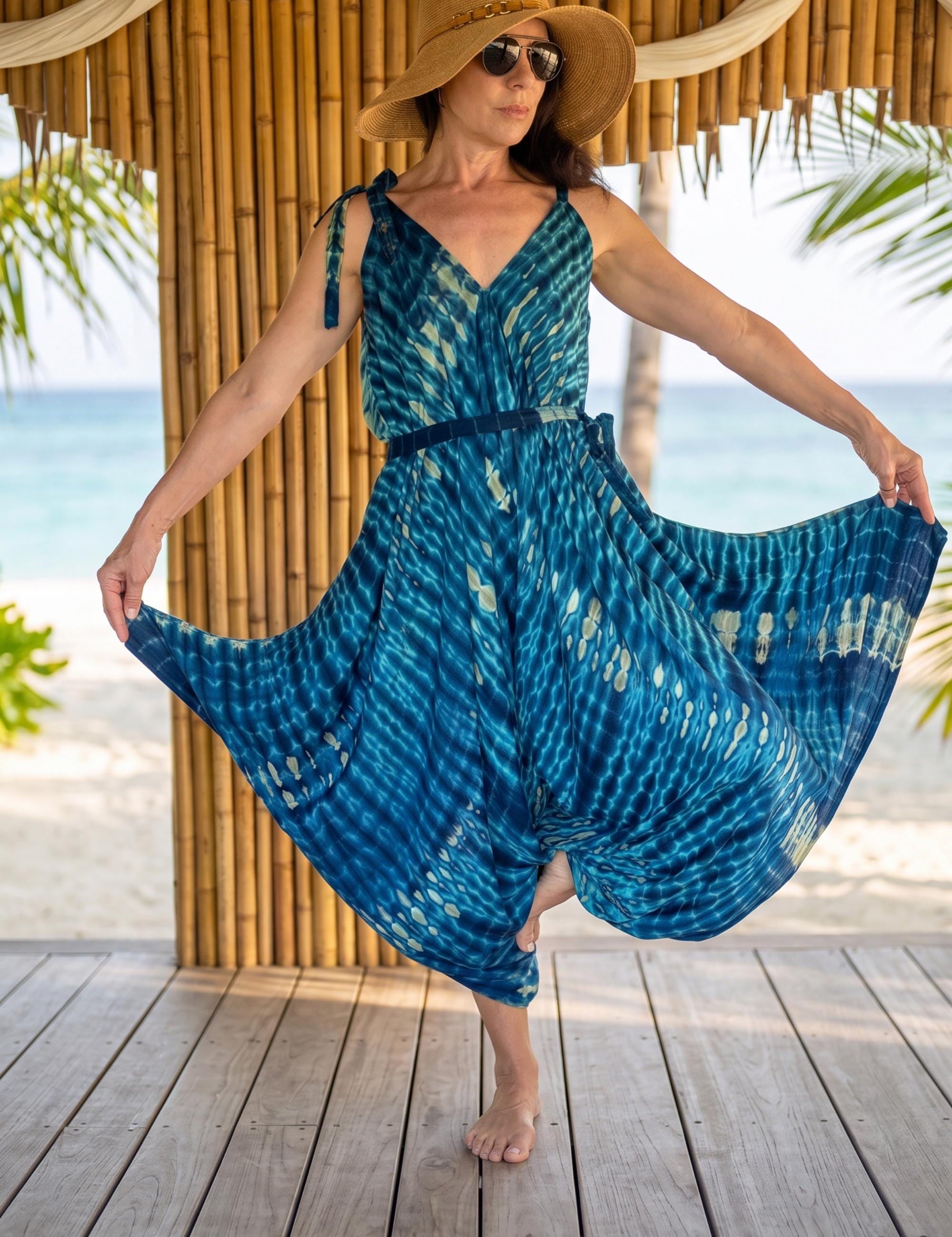 A woman in a blue and white tie-dye dress and a wide-brimmed hat stands on a wooden deck, with palm trees and the ocean visible in the background.