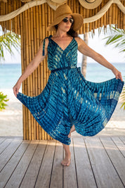 A woman in a blue and white tie-dye dress and a wide-brimmed hat stands on a wooden deck, with palm trees and the ocean visible in the background.