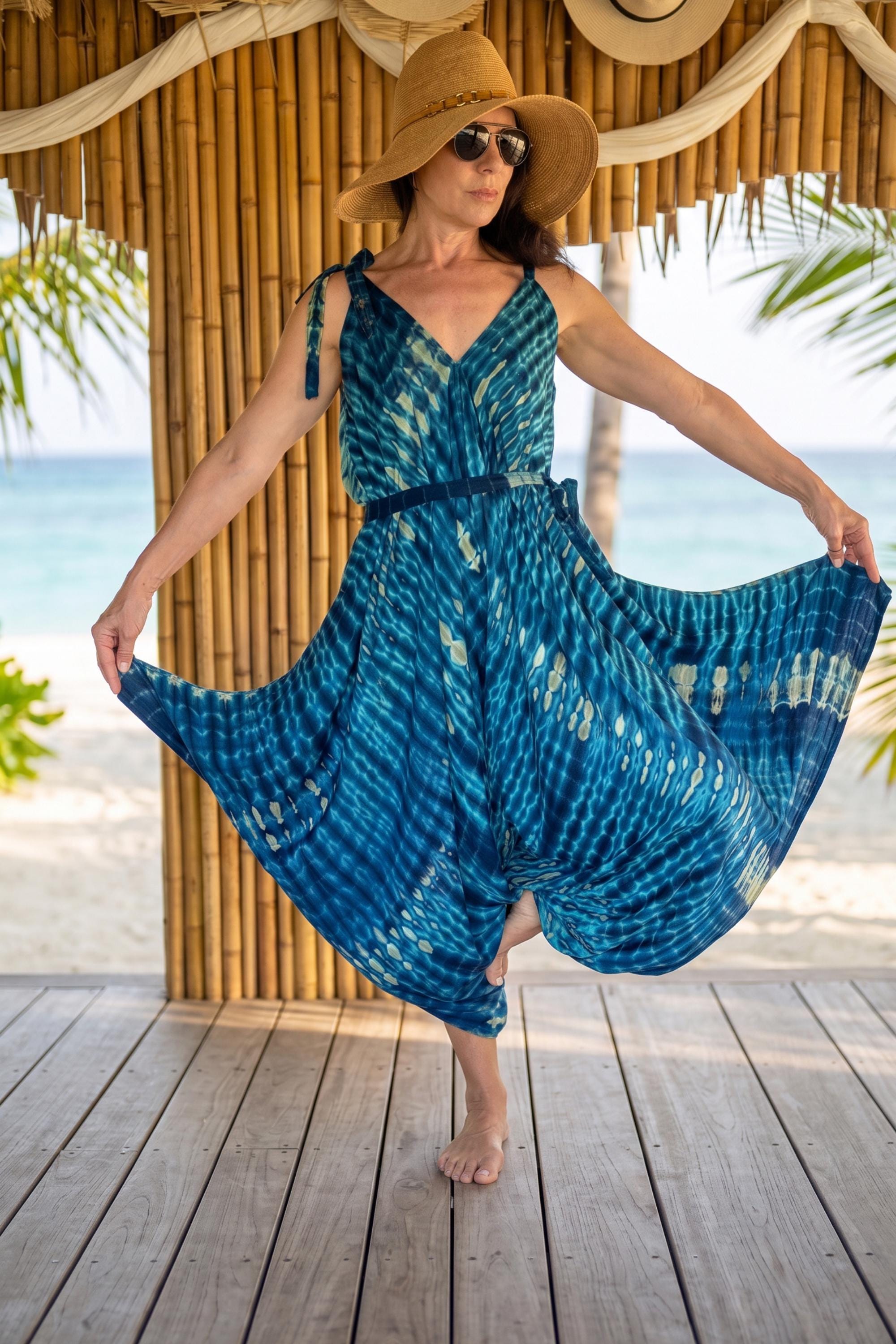 A woman in a blue and white tie-dye dress and a wide-brimmed hat stands on a wooden deck, with palm trees and the ocean visible in the background.