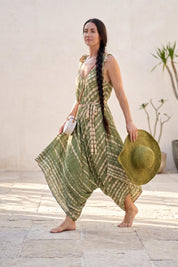 A woman in a green and white tie-dye dress and a wide-brimmed hat walks on a tiled floor, carrying a white purse.