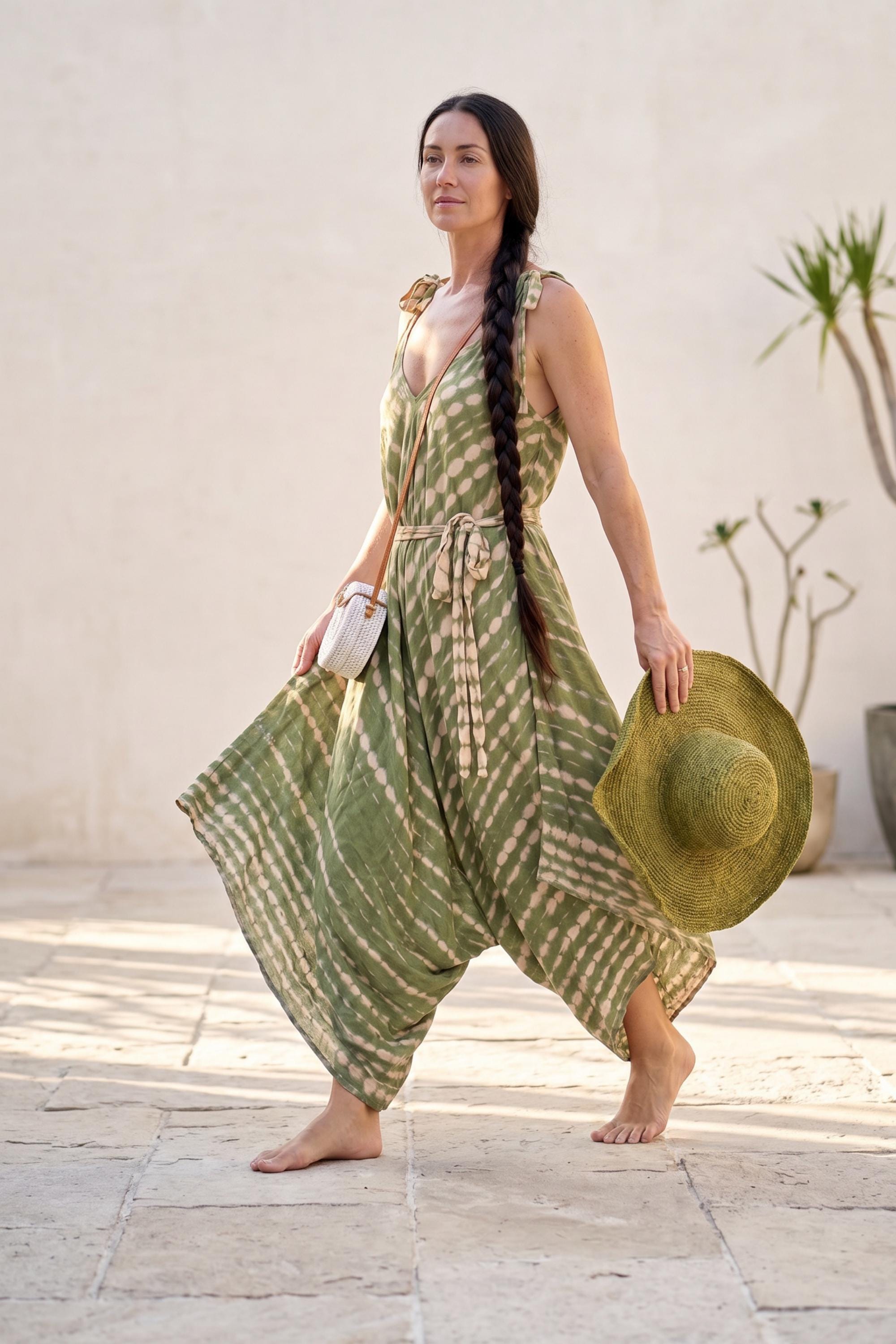 A woman in a green and white tie-dye dress and a wide-brimmed hat walks on a tiled floor, carrying a white purse.