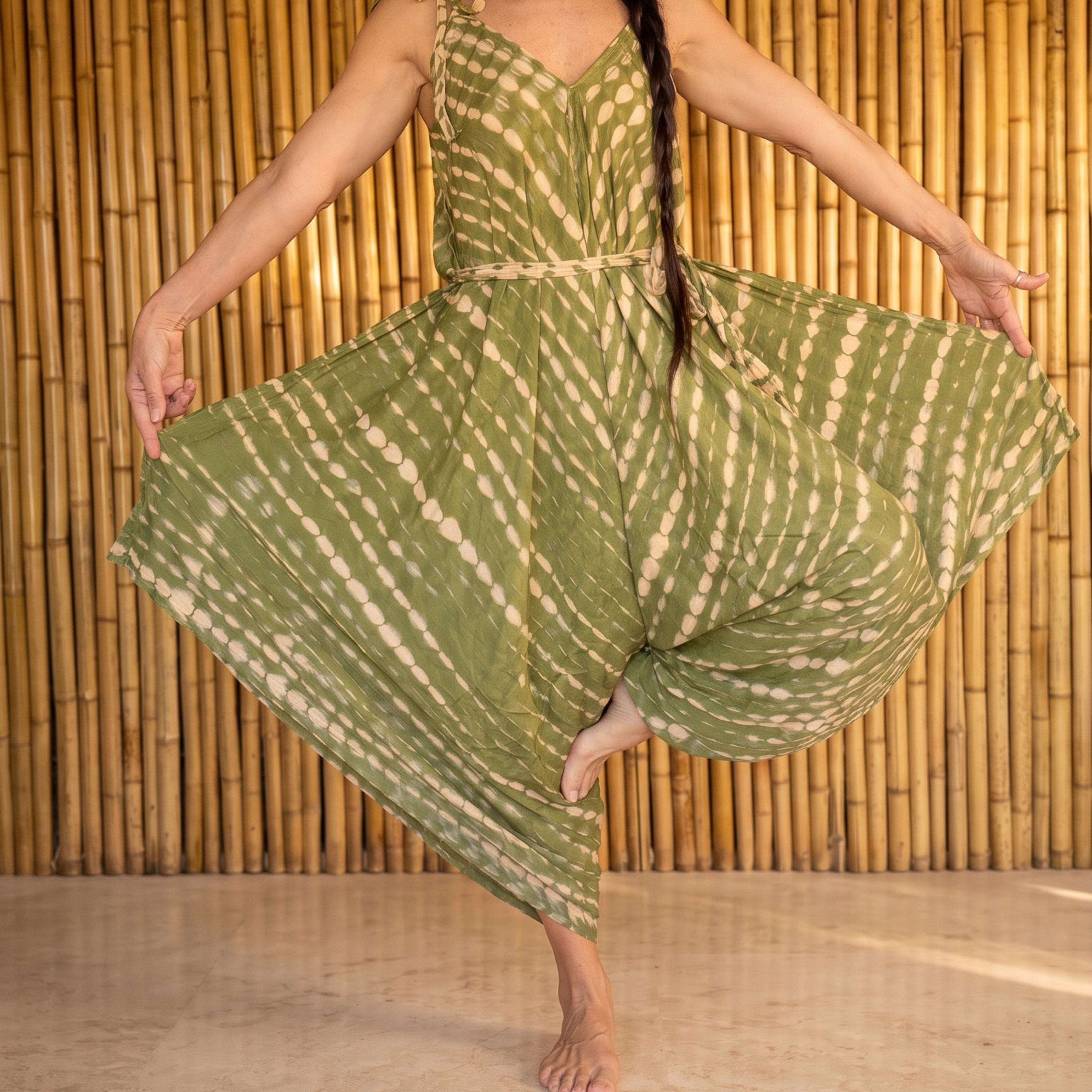 A woman in a green and white patterned dress is performing a yoga pose, with her arms outstretched and her feet balanced on one leg.