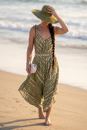 A woman in a green and white patterned dress walks along the beach, wearing a wide-brimmed hat and carrying a white purse.