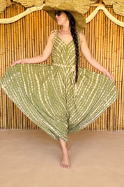 A woman wearing a green and white tie-dye dress and a wide-brimmed hat stands in front of a bamboo wall, with her arms outstretched.