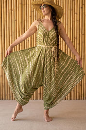 A woman wearing a green and white tie-dye jumpsuit and a wide-brimmed hat stands in front of a bamboo wall, striking a playful pose with her arms outstretched.
