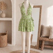 A mannequin wearing a green floral print dress with a halter neckline and a wrap-style waistband, standing in front of a mirror and a wooden bench.