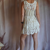 A woman wearing a white crocheted dress with a lace-like pattern, standing against a pink background.