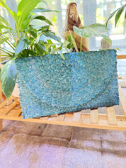 A blue woven purse is placed on a wooden surface, surrounded by lush green plants.