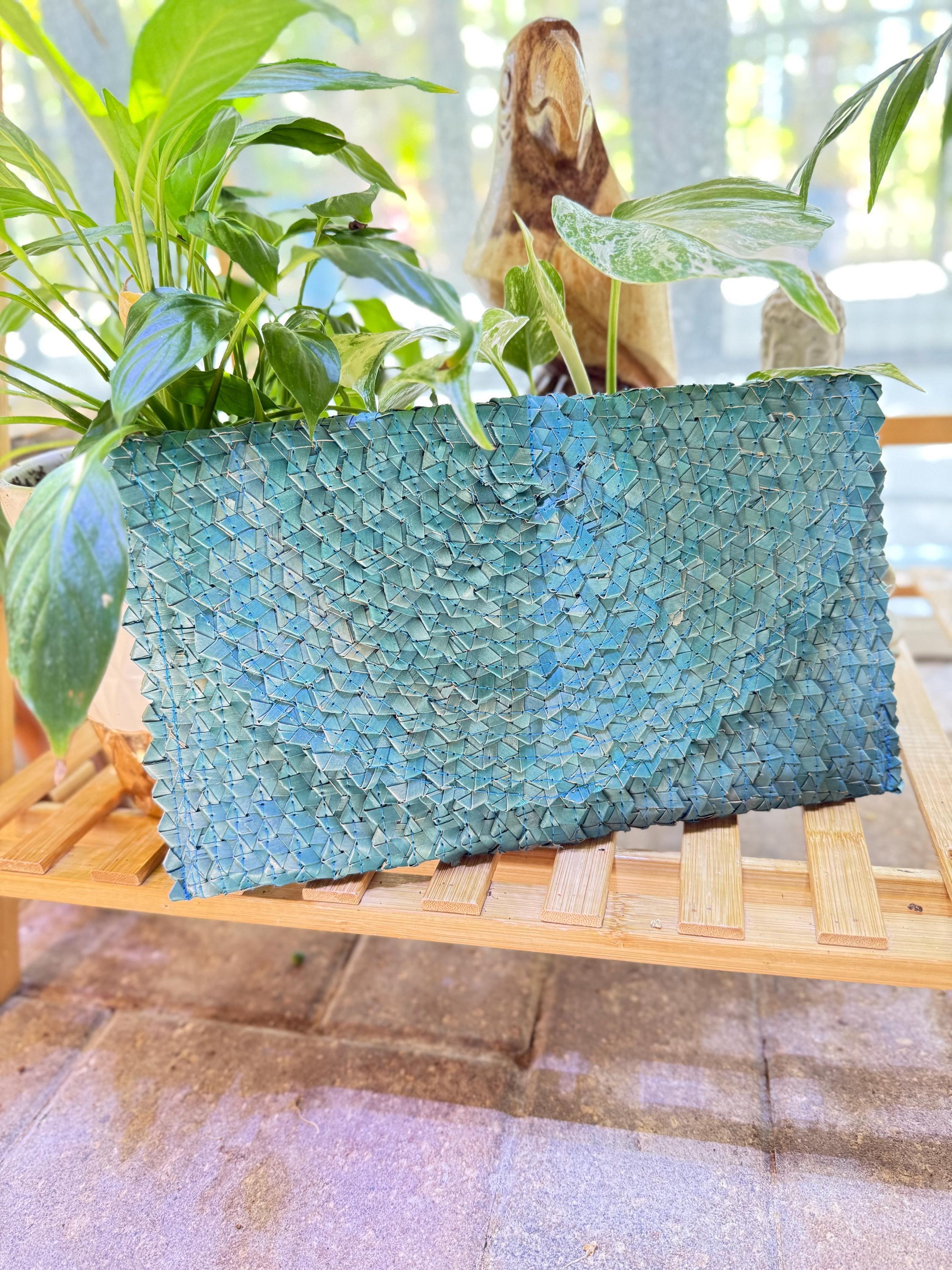 A blue woven purse is placed on a wooden surface, surrounded by lush green plants.