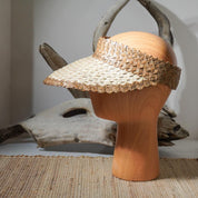 a wooden headless mannequin wearing a straw hat with a woven pattern. The hat is placed on a woven mat, and there is a wooden log or branch in the background.