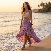 A woman in a white lace top and a purple floral skirt walks along the beach during sunset, with palm trees and the ocean in the background.