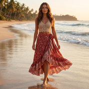 A woman in a red and white floral skirt and a beige crop top walks along a beach at sunset, with palm trees and the ocean in the background.