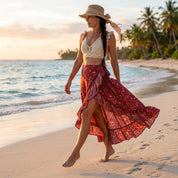 A woman in a white top and red skirt walks along a sandy beach, with palm trees and a sunset in the background.