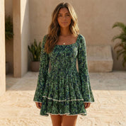 A young woman wearing a green and white floral print dress with long sleeves and a fitted bodice, standing in an outdoor setting with a stone wall and potted plants in the background.