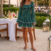 A woman in a green floral dress stands in front of a table with a white tablecloth, candles, and flowers, suggesting a romantic or intimate setting.