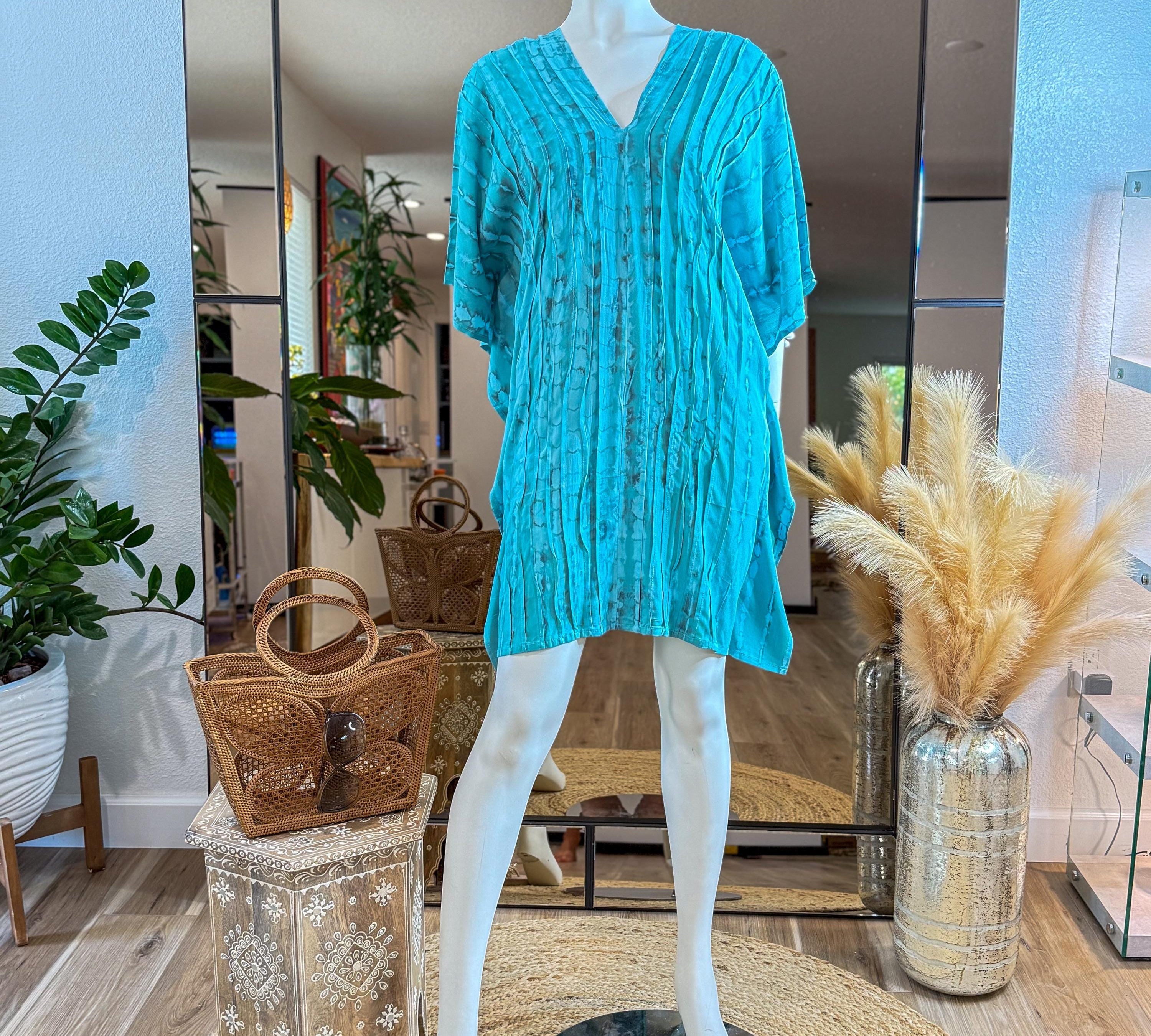 A mannequin wearing a turquoise dress with a ruched neckline and short sleeves, placed on a wooden stand with a woven handbag. The room has a mirror and some potted plants, creating a stylish and inviting atmosphere.