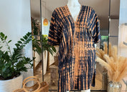 a mannequin wearing a tie-dye dress with a pattern of orange and black stripes. The dress is displayed in a store setting, with potted plants and a mirror in the background.