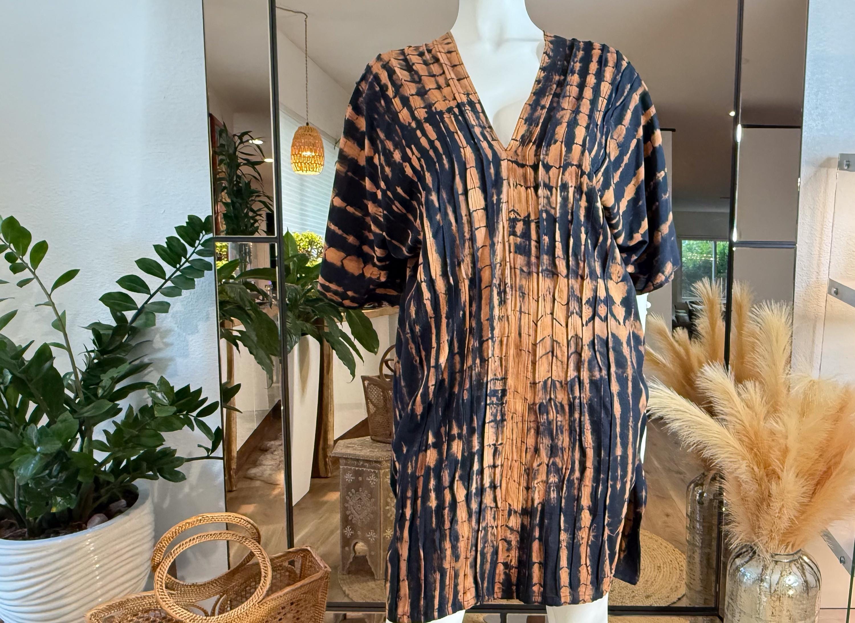 a mannequin wearing a tie-dye dress with a pattern of orange and black stripes. The dress is displayed in a store setting, with potted plants and a mirror in the background.