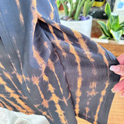 a close-up view of a fabric with a tie-dye pattern, with a person's hand holding the fabric. In the background, there are potted plants, adding a natural element to the scene.