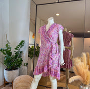 A mannequin wearing a pink floral wrap dress is displayed in a clothing store, with a large mirror behind it. The dress has a ruffled neckline and a wrap-style skirt. The store also features potted plants and decorative items, creating a pleasant and inviting atmosphere.