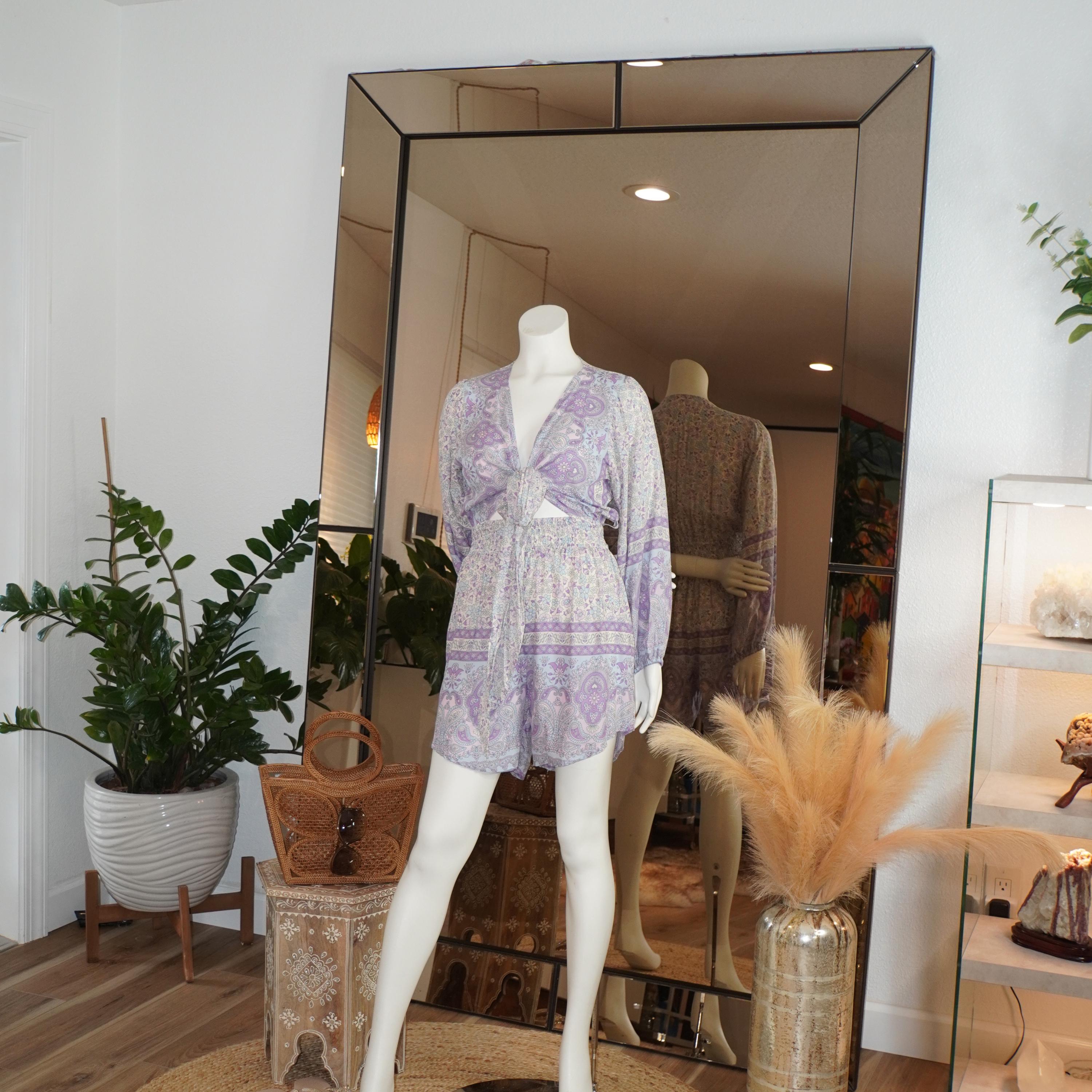 a mannequin wearing a purple and white patterned dress, standing in front of a large mirror. The room has a white color scheme, with a large mirror on the wall and a potted plant nearby.