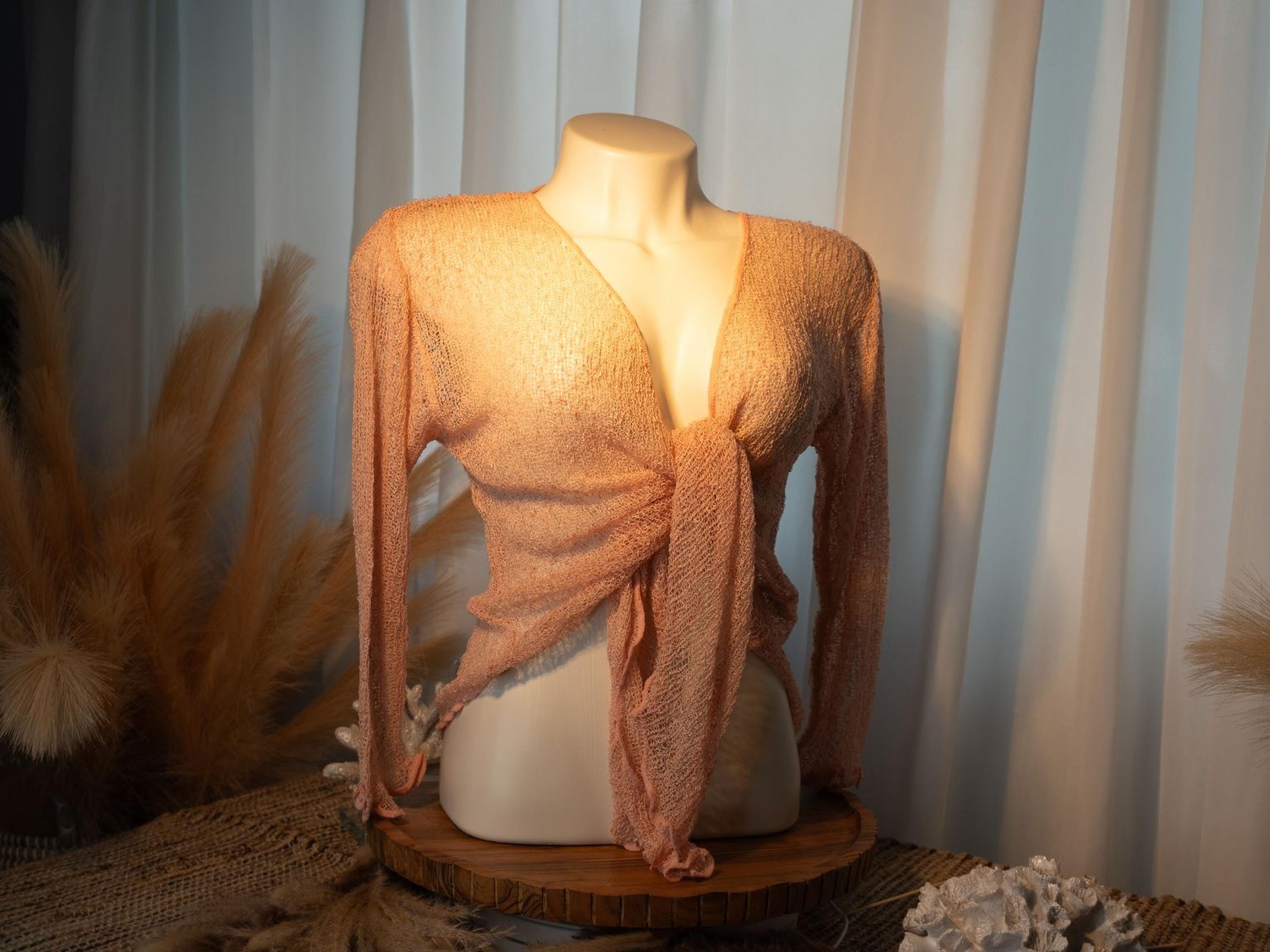 A mannequin wearing a peach-colored knitted top with long sleeves, displayed on a wooden stand against a white curtain background.