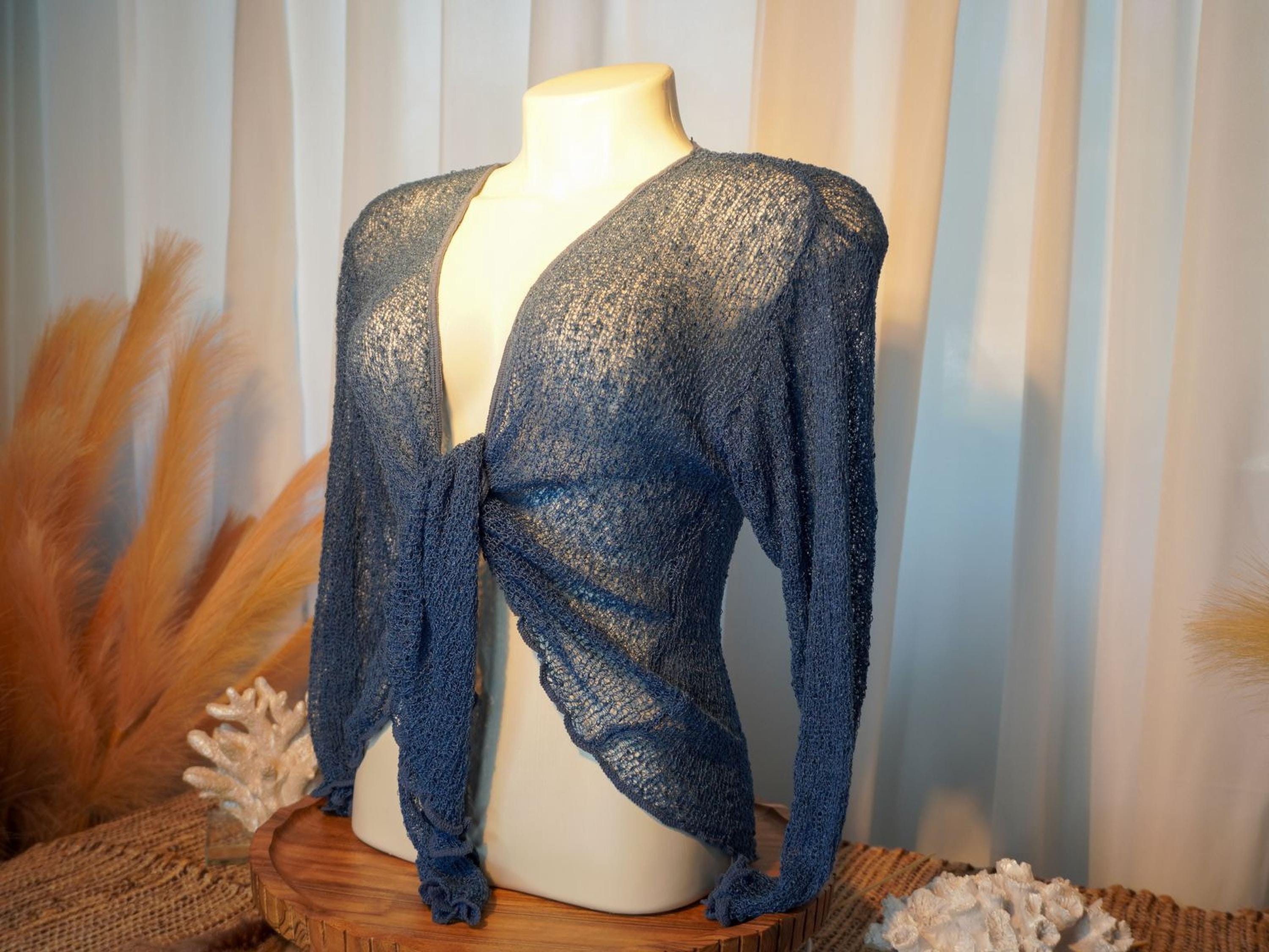 A mannequin wearing a blue knitted top with long sleeves, displayed on a wooden stand with coral and feathers in the background.