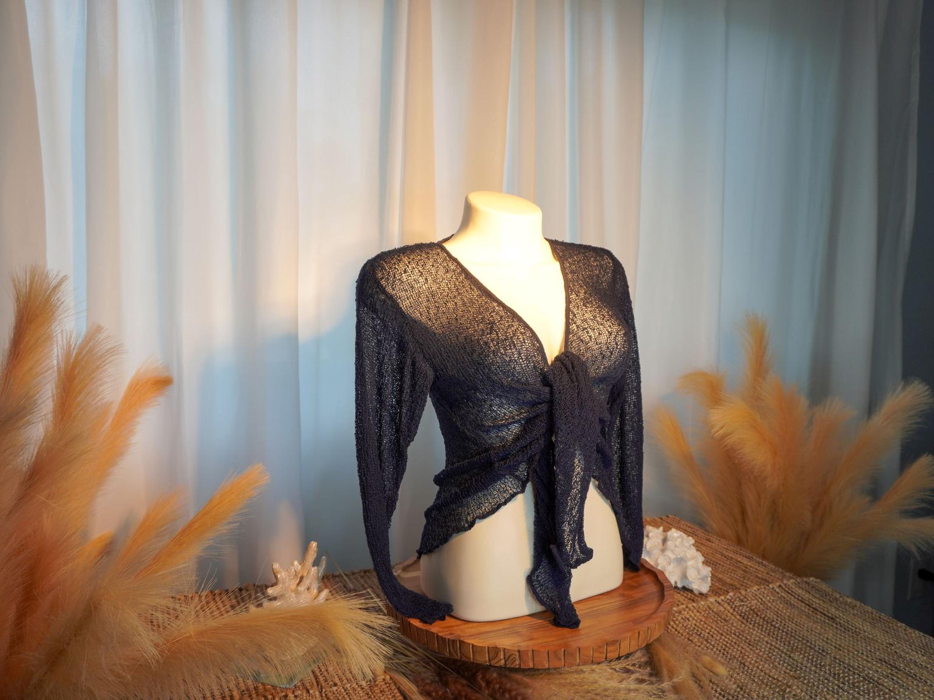 A mannequin wearing a black lace top with long sleeves, placed on a wooden stand with a woven mat and some orange feathers.