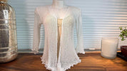 a white knitted cardigan displayed on a mannequin, placed on a wooden surface. Next to the cardigan, there is a candle and a vase, creating a cozy and inviting atmosphere.