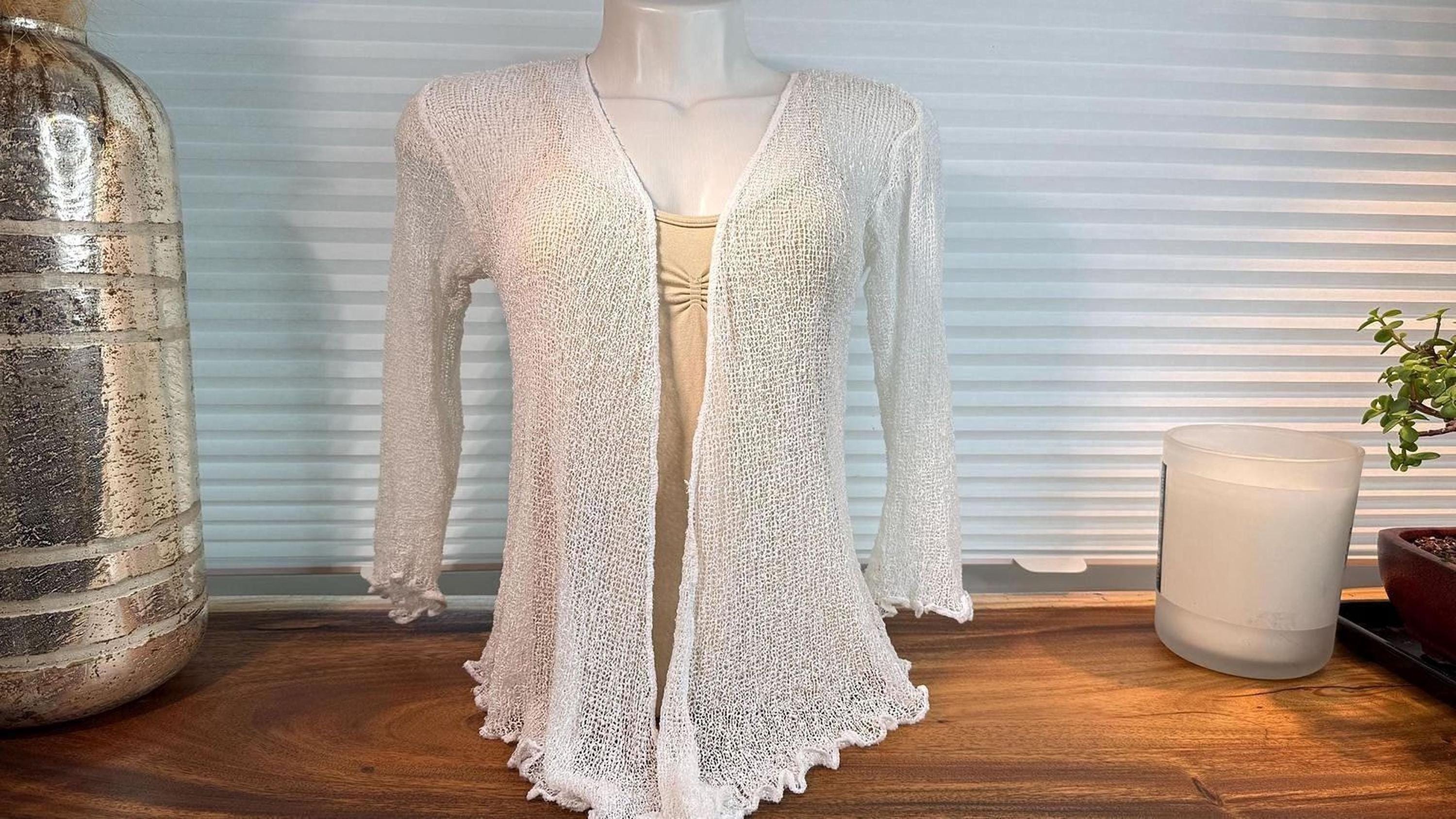 a white knitted cardigan displayed on a mannequin, placed on a wooden surface. Next to the cardigan, there is a candle and a vase, creating a cozy and inviting atmosphere.