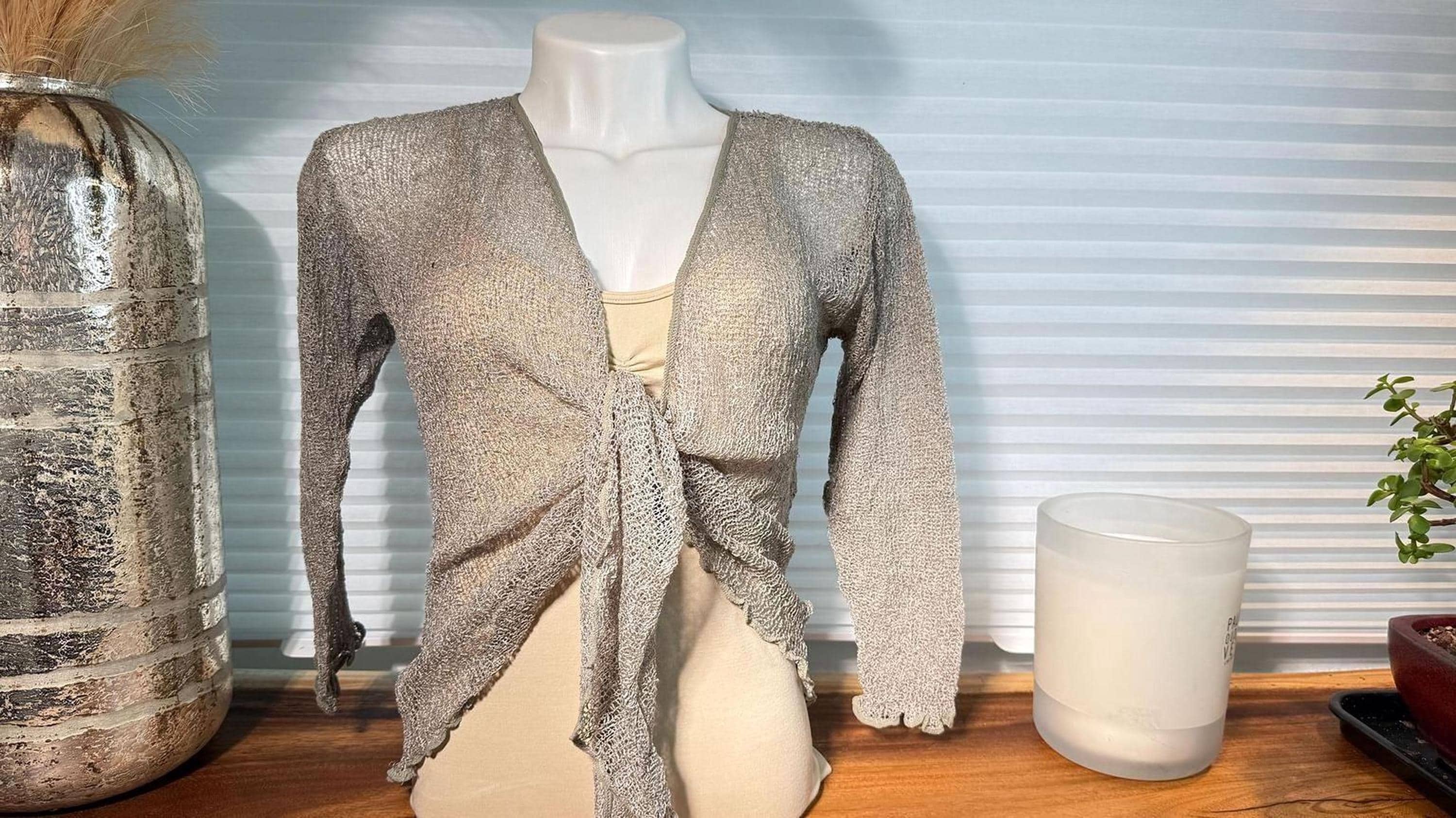 a mannequin wearing a gray knitted cardigan, placed on a wooden surface next to a candle and a decorative vase.