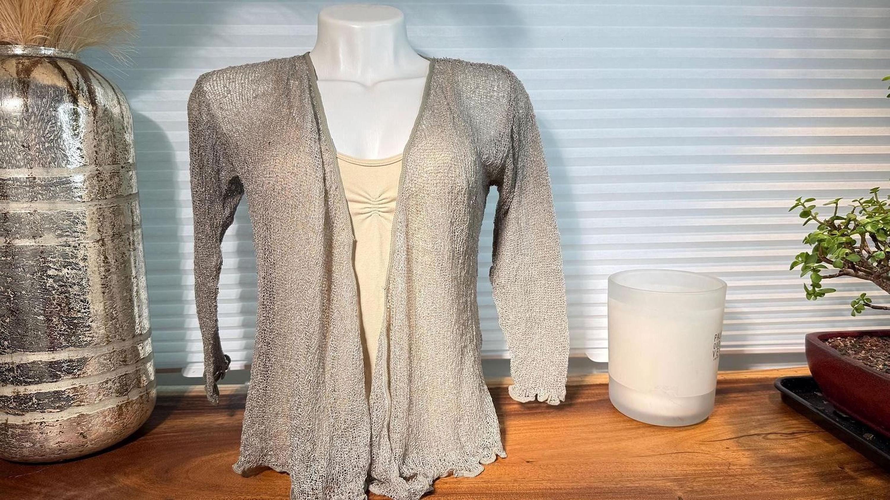 a mannequin wearing a beige sweater with long sleeves, placed on a wooden surface next to a silver vase and a white candle.