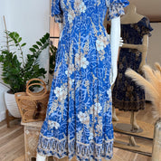 A mannequin wearing a blue floral print dress with a ruffled collar and sleeves, placed in a room with potted plants and a mirror.