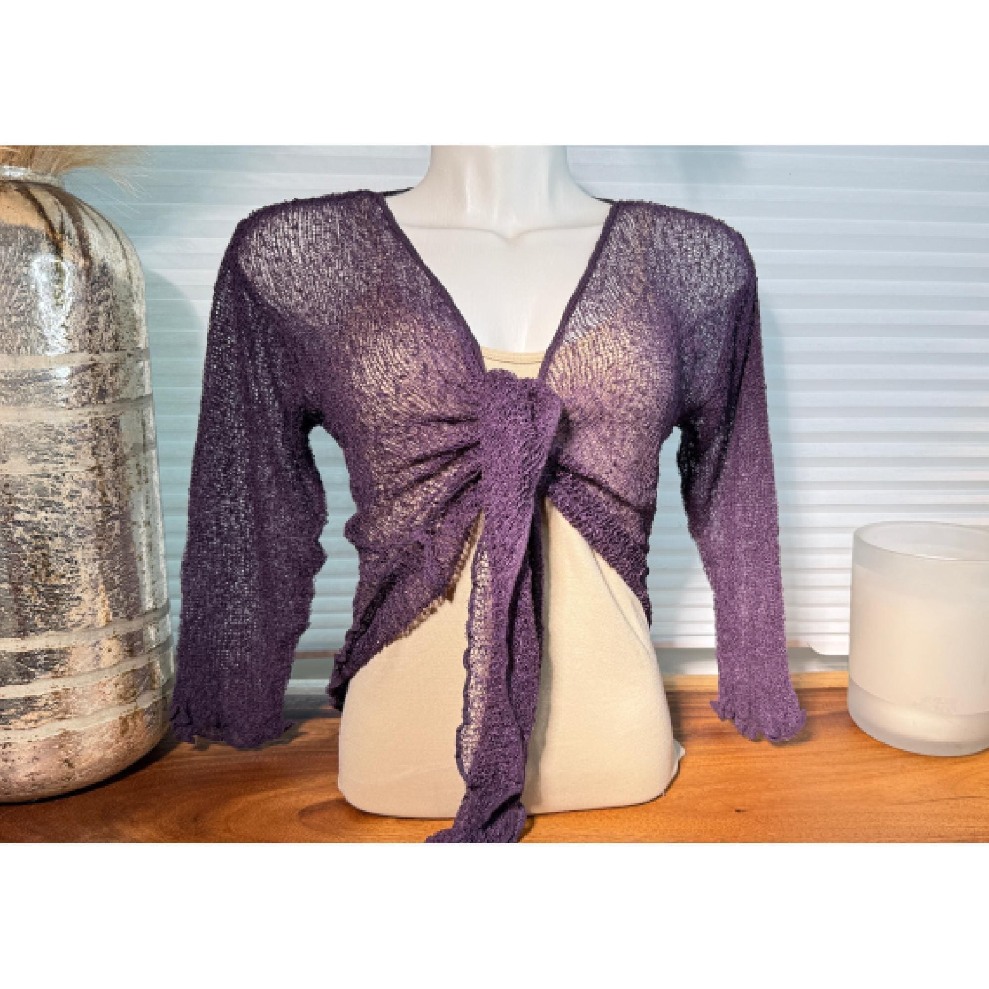 A mannequin wearing a purple knitted wrap top with a tie detail, placed on a wooden surface next to a glass container and a candle.
